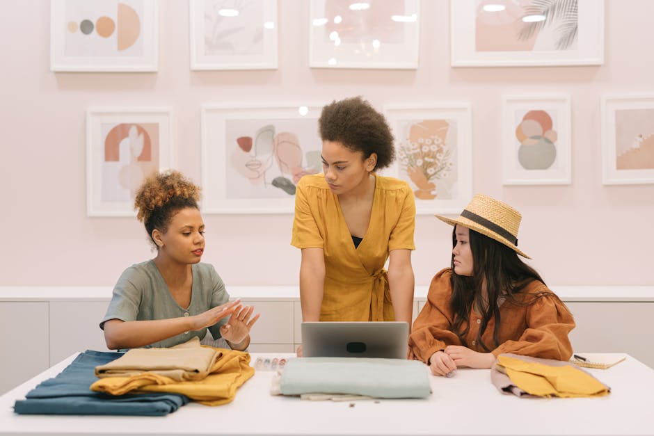 Three women collaborating over design ideas in a fashion studio with fabric samples and art on the walls.